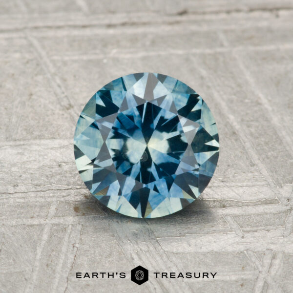 1.27-Carat Montana Sapphire (Heated) A 1.27-carat heated Montana Sapphire, round brilliant-cut and blue, is displayed on a textured gray surface with "Earth's Treasury" at the bottom.