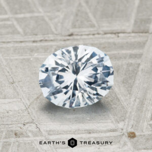 A 1.11-Carat Montana Sapphire gemstone glimmers on a textured gray surface, with “EARTH’S TREASURY” and a hexagonal logo beneath.
