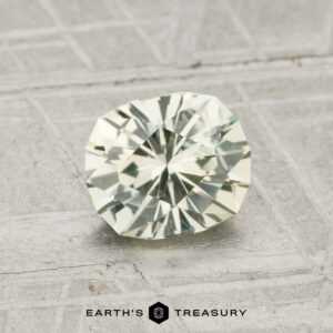 A 1.25-Carat Montana Sapphire, light green and faceted, rests on a textured gray surface with "EARTH'S TREASURY" displayed at the bottom.