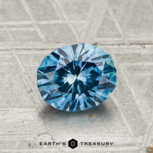 A 1.30-carat heated Montana sapphire with a faceted blue hue rests on a textured metallic surface, with "EARTH'S TREASURY" displayed at the bottom.