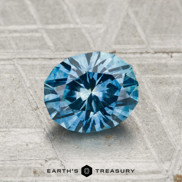 1.30-Carat Montana Sapphire (Heated) A 1.30-carat heated Montana sapphire with a faceted blue hue rests on a textured metallic surface, with "EARTH'S TREASURY" displayed at the bottom.