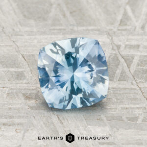 A 1.23-carat cushion-cut Montana sapphire (heated) displayed on a textured gray surface, with the text "EARTH'S TREASURY" below.