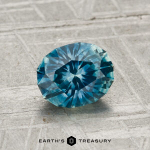 A 1.30-carat heated Montana sapphire, round and faceted, is displayed on a textured gray surface with the "Earth’s Treasury" logo beneath it.