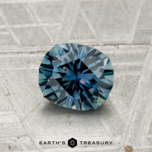A faceted 1.18-carat heated Montana sapphire with a radiating pattern rests on a textured gray surface. The "EARTH'S TREASURY" logo appears at the bottom.