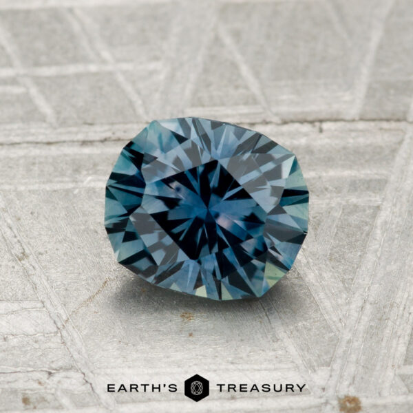 1.18-Carat Montana Sapphire (Heated) A faceted 1.18-carat heated Montana sapphire with a radiating pattern rests on a textured gray surface. The "EARTH'S TREASURY" logo appears at the bottom.