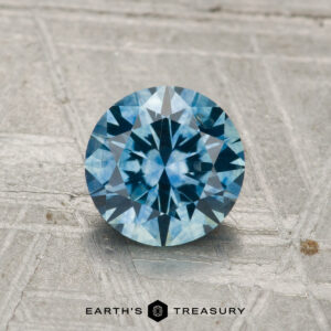 A 1.26-carat heated Montana sapphire, round brilliant-cut and blue, stands upright on a textured gray surface with "EARTH'S TREASURY" displayed below.