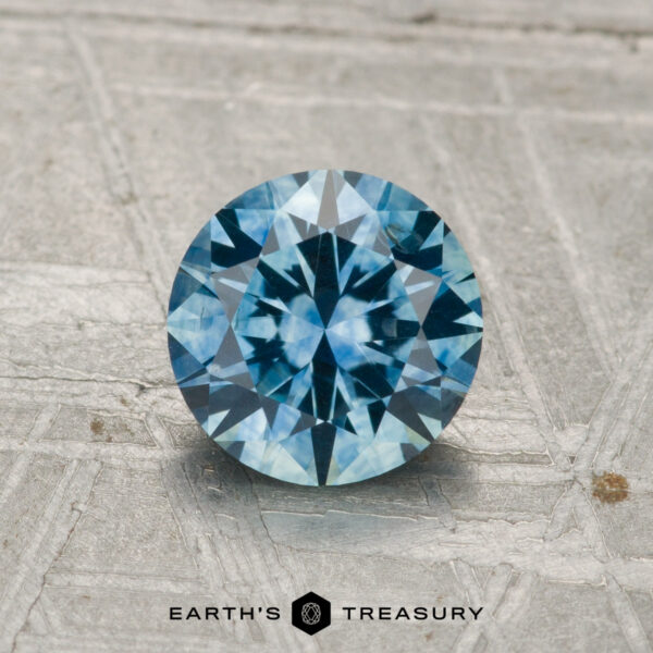 1.26-Carat Montana Sapphire (Heated) A 1.26-carat heated Montana sapphire, round brilliant-cut and blue, stands upright on a textured gray surface with "EARTH'S TREASURY" displayed below.