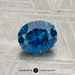 A 1.29-carat heated Montana sapphire with intricate facets is displayed on a textured gray surface. The words "EARTH'S TREASURY" appear at the bottom.