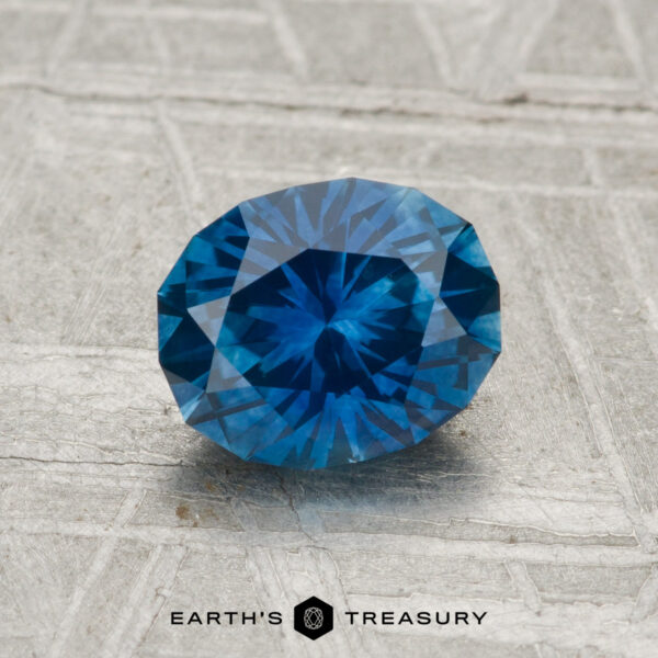 1.29-Carat Montana Sapphire (Heated) A 1.29-carat heated Montana sapphire with intricate facets is displayed on a textured gray surface. The words "EARTH'S TREASURY" appear at the bottom.
