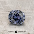 A faceted 1.18-carat Montana Sapphire (heated) in steely blue is displayed on a light stone surface, with “EARTH’S TREASURY” text and logo at the bottom.