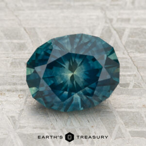 A 2.92-carat heated Montana Sapphire with a faceted oval blue-green cut is showcased on a textured gray surface, featuring the text "Earth's Treasury" below.