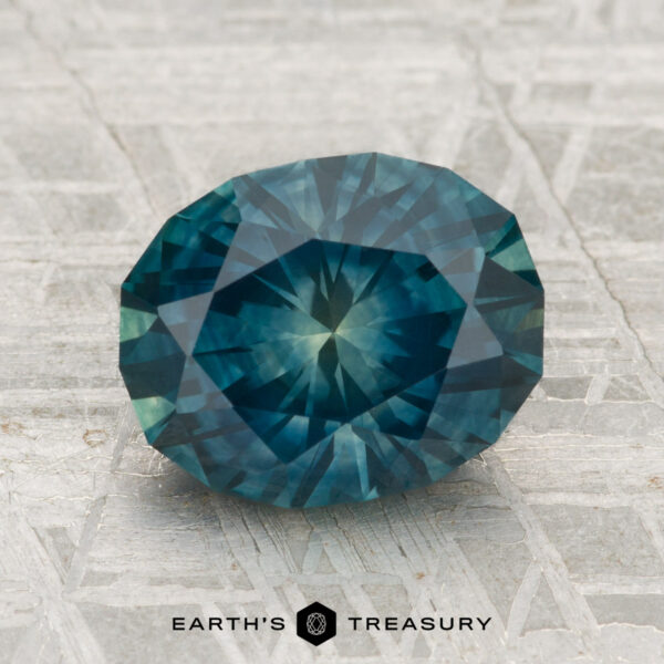 2.92-Carat Montana Sapphire (Heated) A 2.92-carat heated Montana Sapphire with a faceted oval blue-green cut is showcased on a textured gray surface, featuring the text "Earth's Treasury" below.