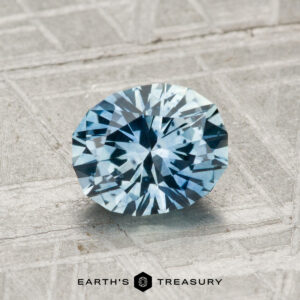 A 1.29-carat heated Montana sapphire, round and blue-green, is displayed on a textured gray surface with the "EARTH'S TREASURY" logo at the bottom.