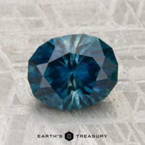 A round, faceted 2.71-carat heated Montana sapphire is displayed on a textured gray surface with "EARTH'S TREASURY" at the bottom.