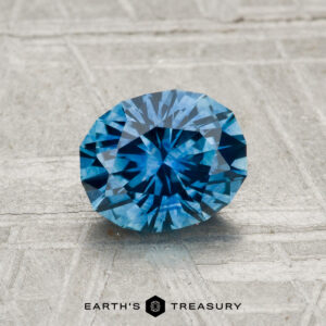 A 1.29-carat heated Montana sapphire, oval and blue, displayed on a textured gray surface with "EARTH'S TREASURY" at the bottom of the image.