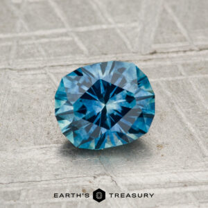 A 1.17-carat heated Montana sapphire with a faceted blue brilliance rests on a textured gray surface, "EARTH'S TREASURY" displayed at the bottom of the image.
