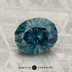 A 2.42-carat heated Montana sapphire, faceted in an oval shape with a teal hue, displayed on a textured light surface, with "EARTH'S TREASURY" beneath.