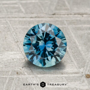 A 1.22-carat, round, brilliant-cut Montana sapphire (heated) is displayed on a textured gray surface with "EARTH'S TREASURY" beneath it.