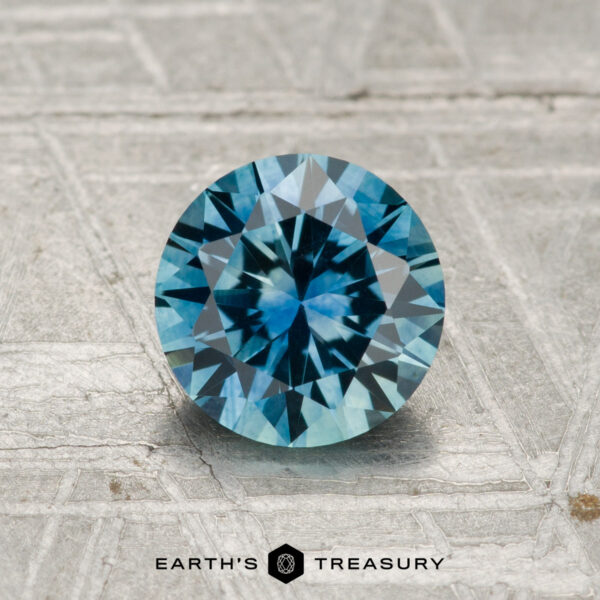 1.22-Carat Montana Sapphire (Heated) A 1.22-carat, round, brilliant-cut Montana sapphire (heated) is displayed on a textured gray surface with "EARTH'S TREASURY" beneath it.