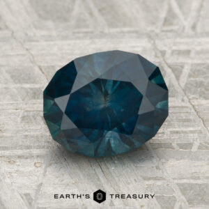 A 2.42-carat heated Montana sapphire, oval and blue-green, is displayed on a textured gray surface with "EARTH'S TREASURY" at the bottom.