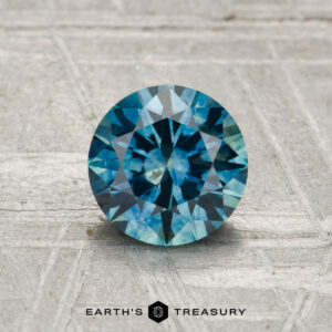1.21-Carat Montana Sapphire (Heated), round brilliant-cut blue gemstone, displayed on a textured gray surface with the "Earth's Treasury" logo at the bottom.