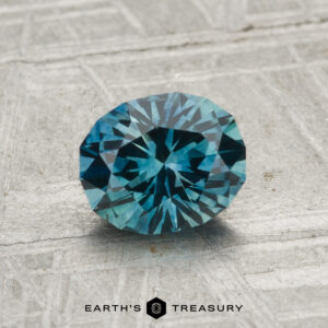 A 1.26-carat heated Montana sapphire, faceted in an oval shape with a blue-green hue, is displayed on a textured gray surface with the text "EARTH'S TREASURY" at the bottom.