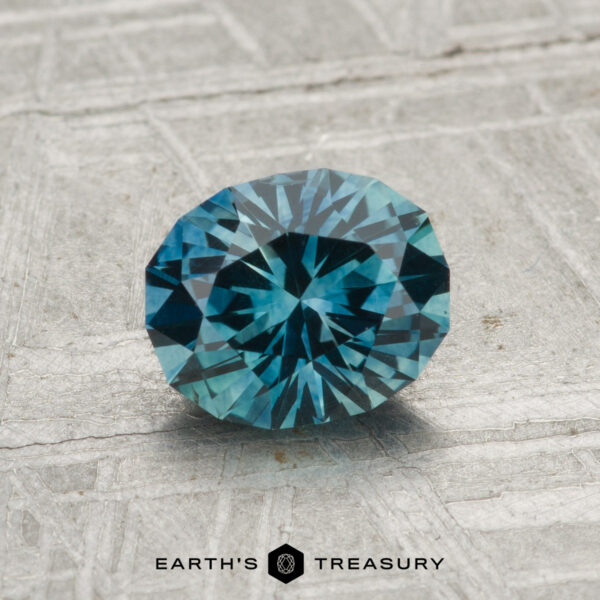 1.26-Carat Montana Sapphire (Heated) A 1.26-carat heated Montana sapphire, faceted in an oval shape with a blue-green hue, is displayed on a textured gray surface with the text "EARTH'S TREASURY" at the bottom.