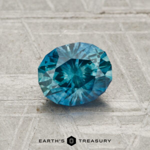 A 1.14 carat heated Montana sapphire with an oval faceted cut is showcased on a textured gray surface, featuring the “Earth’s Treasury” logo at the bottom.