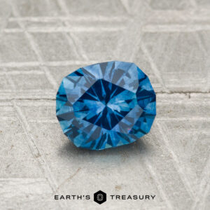 A 1.10-carat heated Montana sapphire rests on a textured gray surface, with "Earth's Treasury" visible at the bottom.