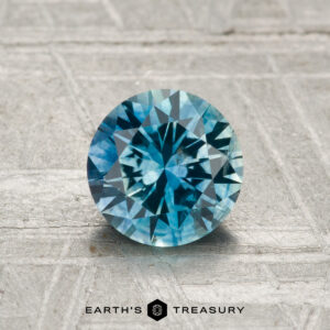 A 1.21-carat faceted Montana sapphire (heated), round and blue-green, displayed on a textured gray surface with "EARTH'S TREASURY" at the bottom.