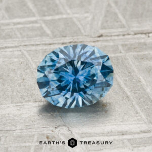 A round, faceted 1.26-carat heated Montana sapphire with a blue hue rests on a textured grey surface, featuring the "EARTH'S TREASURY" logo and text at the bottom.