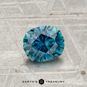 A 1.08-carat heated Montana sapphire with teal-blue highlights rests on a patterned metallic surface, with the text "Earth's Treasury" at the bottom.