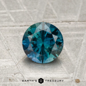 A 1.20-carat heated Montana sapphire, round and faceted in blue-green hues, is displayed on a textured gray surface with "Earth’s Treasury" at the bottom.
