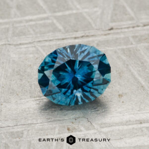 A 1.25-carat heated Montana sapphire, faceted in an oval shape, is shown on a textured gray surface with "EARTH'S TREASURY" below.