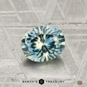 A 1.14 carat heated Montana sapphire with blue-green hues rests on a textured metallic surface. The text "EARTH'S TREASURY" is displayed at the bottom of the image.