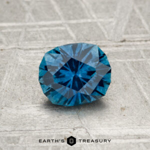 A 1.04-carat heated Montana sapphire with a blue, faceted oval shape rests on a textured gray surface. "EARTH'S TREASURY" is displayed at the bottom of the image.