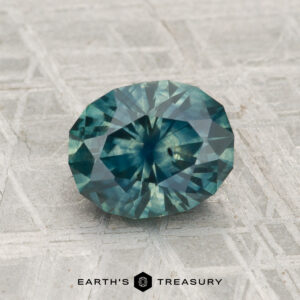 A 1.83-carat heated Montana sapphire in faceted teal-blue rests on a patterned surface with "EARTH'S TREASURY" displayed at the bottom.