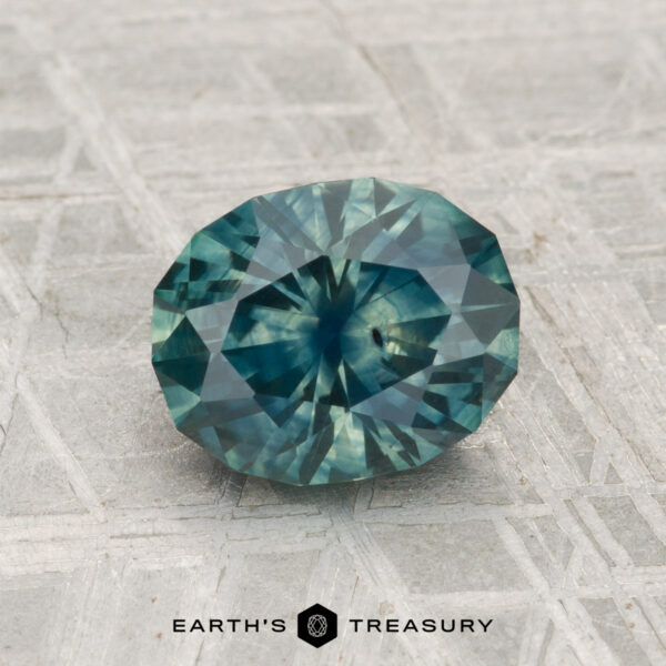 1.83-Carat Montana Sapphire (Heated) A 1.83-carat heated Montana sapphire in faceted teal-blue rests on a patterned surface with "EARTH'S TREASURY" displayed at the bottom.