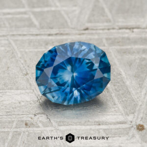 A faceted blue 1.14 Carat Montana Sapphire (Heated) rests on a textured gray surface, with "EARTH'S TREASURY" and a hexagon logo at the bottom.