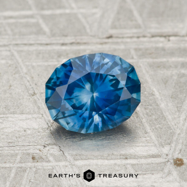 1.14 Carat Montana Sapphire (Heated) A faceted blue 1.14 Carat Montana Sapphire (Heated) rests on a textured gray surface, with "EARTH'S TREASURY" and a hexagon logo at the bottom.