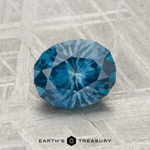 A 1.77-carat oval-cut Montana Sapphire (heated) is showcased on a textured, light surface with "EARTH'S TREASURY" and a hexagonal logo below.