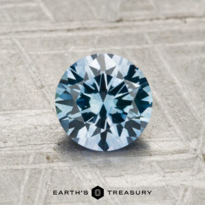 A 1.19-carat heated Montana sapphire, brilliant-cut and blue, displayed on a textured gray surface with the "Earth's Treasury" logo at the bottom.
