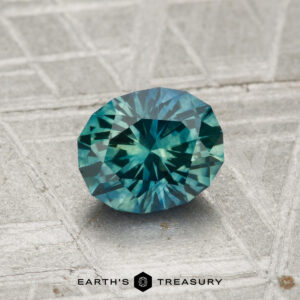 A 1.14-carat heated Montana sapphire, faceted in an oval cut, rests on a textured gray surface with "EARTH'S TREASURY" and a hexagonal logo below.