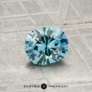 A 1.03-carat heated Montana sapphire with a blue hue rests on a textured metallic surface, with the text "EARTH'S TREASURY" at the bottom of the image.