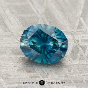 A 1.65-carat Montana Sapphire (heated), oval-cut with a starburst pattern, rests on a textured silver-colored surface. "EARTH'S TREASURY" is visible at the bottom.