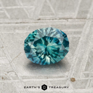 A round-cut 1.13-carat Montana sapphire (heated) in teal rests on a textured metallic surface with the "EARTH'S TREASURY" logo and text below.