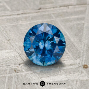 1.17-carat heated Montana sapphire with a round brilliant cut showcased on a textured gray surface, featuring the "Earth's Treasury" text at the bottom.