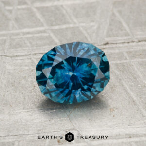 A 1.25-carat heated Montana sapphire with an oval cut is shown on a textured gray surface, featuring the text "EARTH'S TREASURY" and a hexagon logo at the bottom.