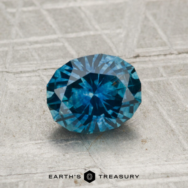 1.25-Carat Montana Sapphire (Heated) A 1.25-carat heated Montana sapphire with an oval cut is shown on a textured gray surface, featuring the text "EARTH'S TREASURY" and a hexagon logo at the bottom.