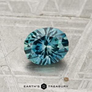 A round, faceted 1.11-carat Montana sapphire (heated) in blue-green hues is displayed on a textured metallic surface with the "EARTH'S TREASURY" text and logo at the bottom.